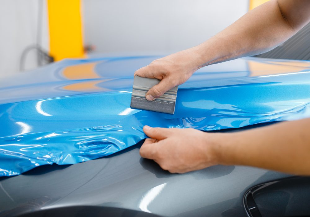 Car wrapping, mechanic with squeegee installs protective vinyl foil or film on vehicle hood. Worker makes auto detailing. Automobile paint protection coating, professional tuning